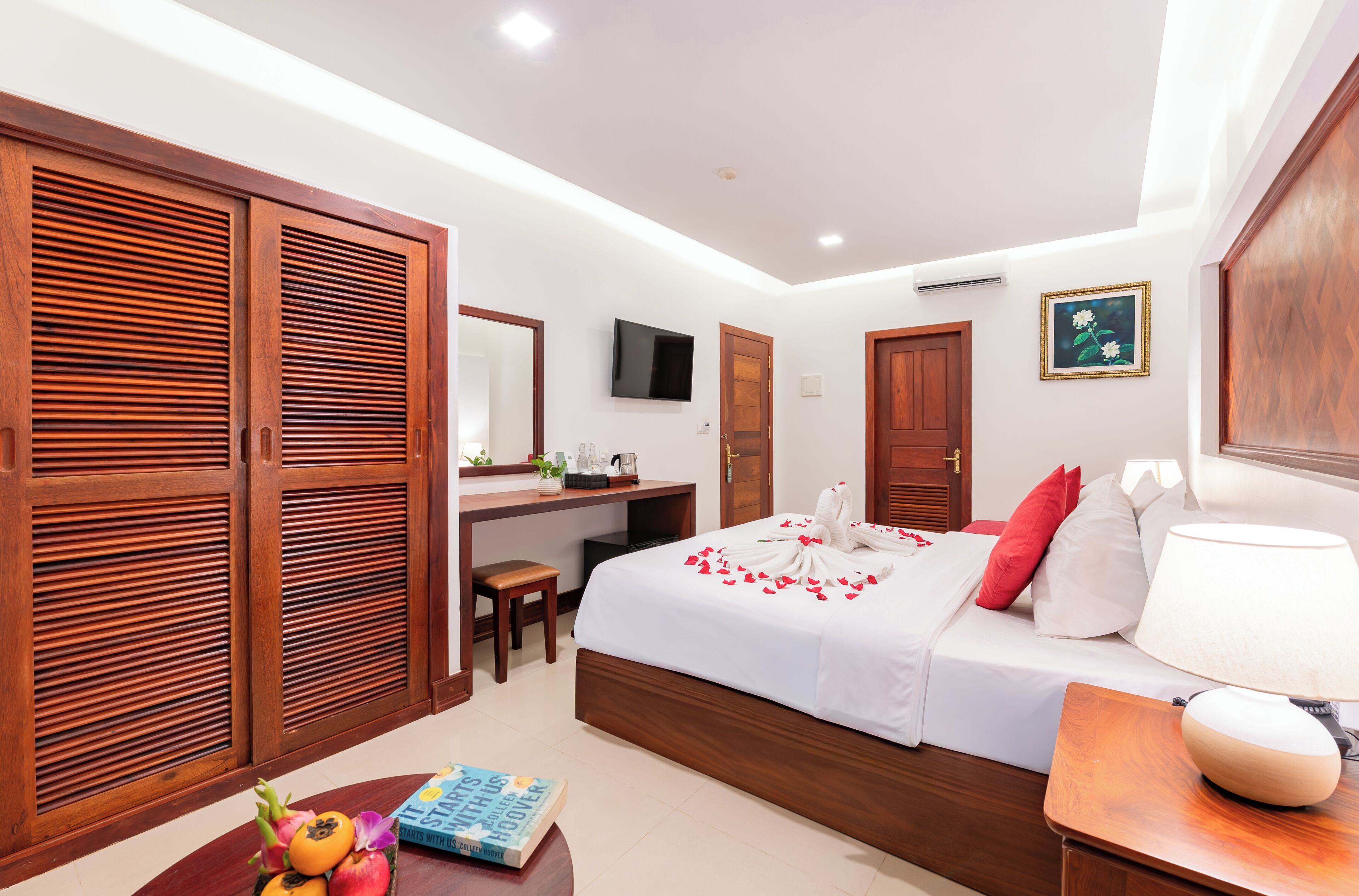 A hotel room with a wooden wardrobe, a desk with a chair, a bed with white bedding and red pillows, a bedside table with a lamp, and a TV on the wall.