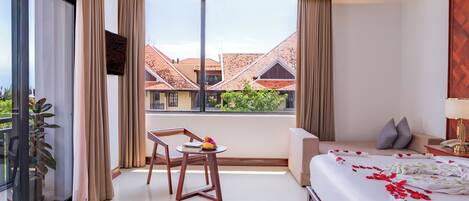 A modern hotel room with a balcony, a bed with rose petals, a small table with fruit, and a view of buildings.
