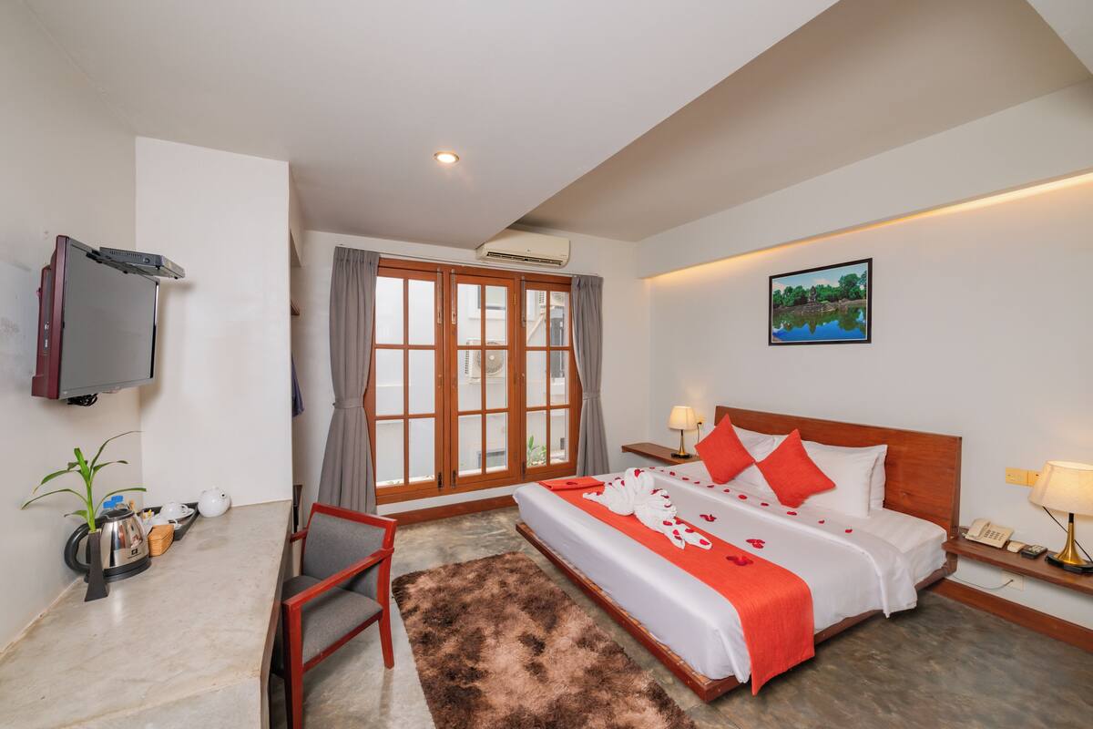 A hotel room with a bed, a television, a desk, a chair, a plant, and a window with curtains.
