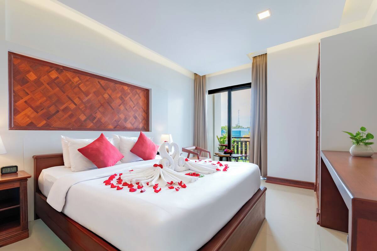 A hotel room with a bed, a nightstand, a digital clock, a large window with a view, and a potted plant.