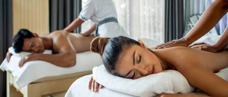 A massage therapy room with two clients receiving treatment, a therapist attending to them, and a window with blinds.