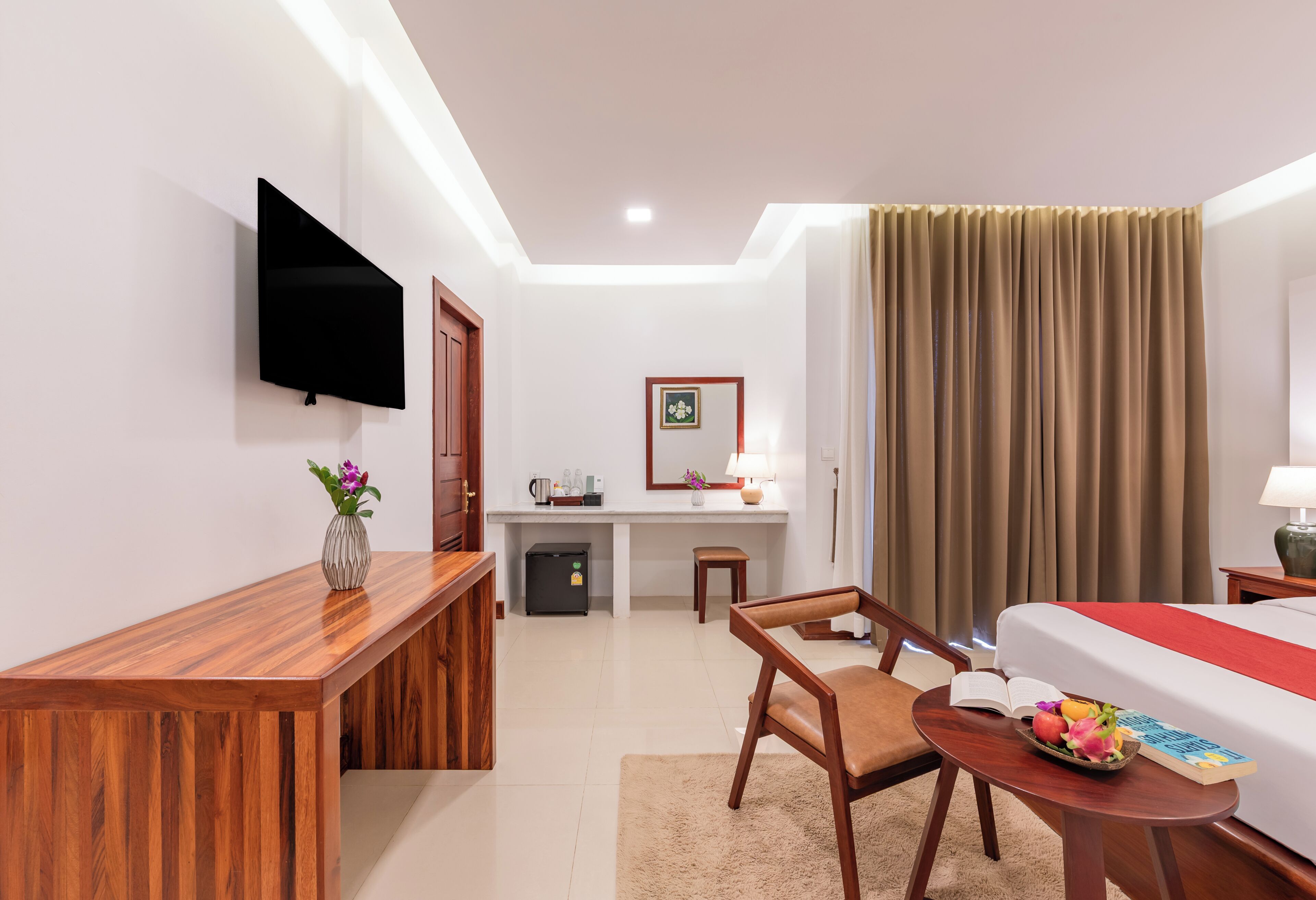 A hotel room with a wooden desk, a television, a bed, a small table with a book and fruit, and a desk with a lamp and picture frame.