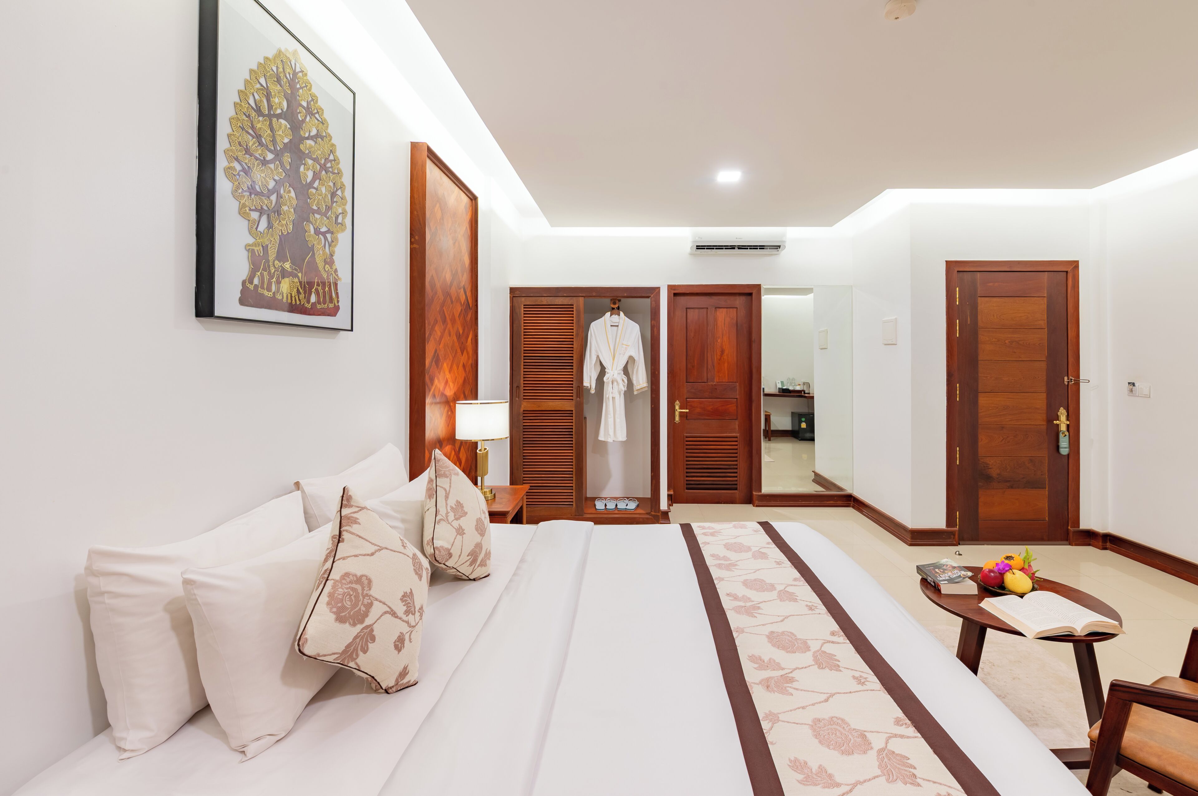 Quarto de hotel com cama, mesa de cabeceira com um livro e frutas, roupão pendurado na porta e quadro na parede.