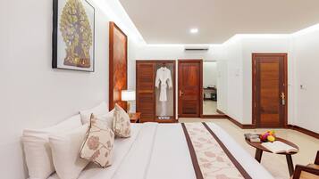 Habitación de hotel con una cama, una mesita de noche con un libro y fruta, una bata colgada en la puerta, y un cuadro en la pared.