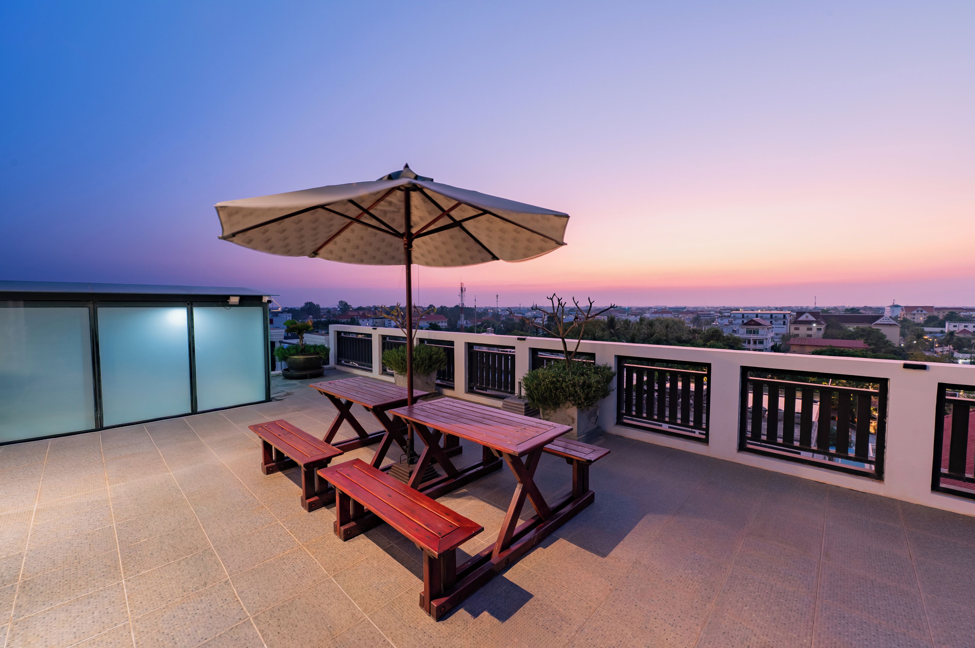 A rooftop terrace with picnic tables and an umbrella, overlooking a cityscape at sunset.
