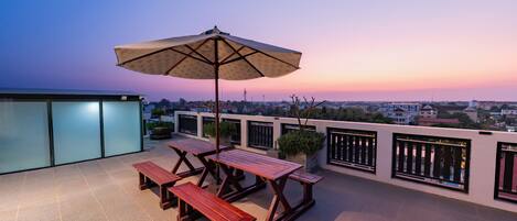 A rooftop terrace with picnic tables and an umbrella, overlooking a cityscape at sunset.