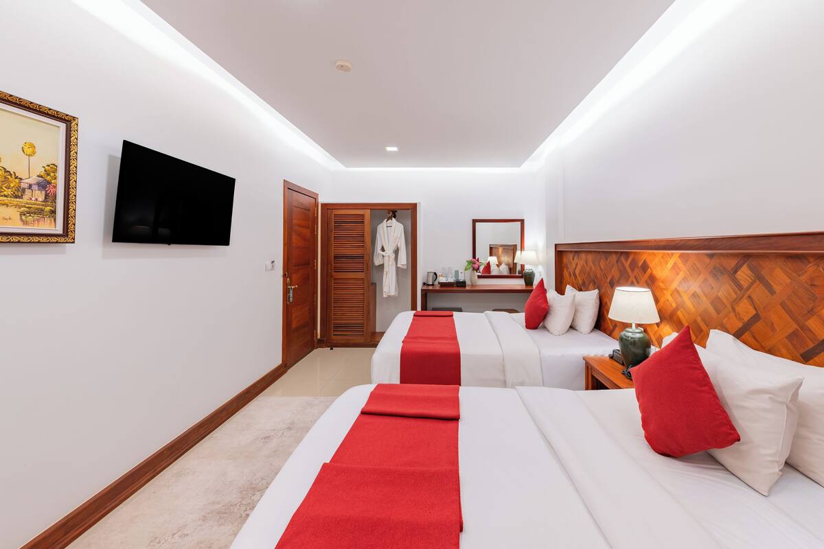 A hotel room with two beds, a TV, a wooden headboard, a mirror, a bedside table with a lamp, and a red runner.