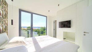 A bedroom with a large bed, a TV mounted on the wall, and a sliding glass door leading to a balcony with a view of greenery and buildings. - Casa Bela with heated pool and luxury of 5 stars (Klimno (Krk))