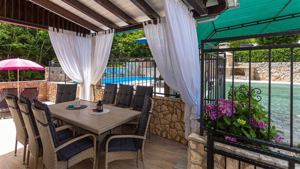 Une terrasse extérieure avec un mur en pierre, des meubles en osier, une table avec une lanterne et une vue sur une piscine.