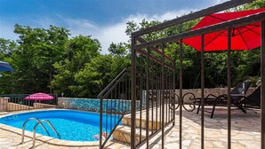 Une piscine avec une terrasse en pierre, un portail en métal et un parasol rouge.