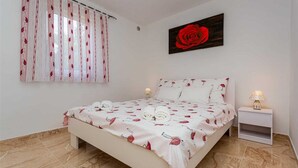 Une chambre à coucher avec un lit, une table de chevet, une lampe, une fenêtre avec des rideaux et un tableau représentant une rose.