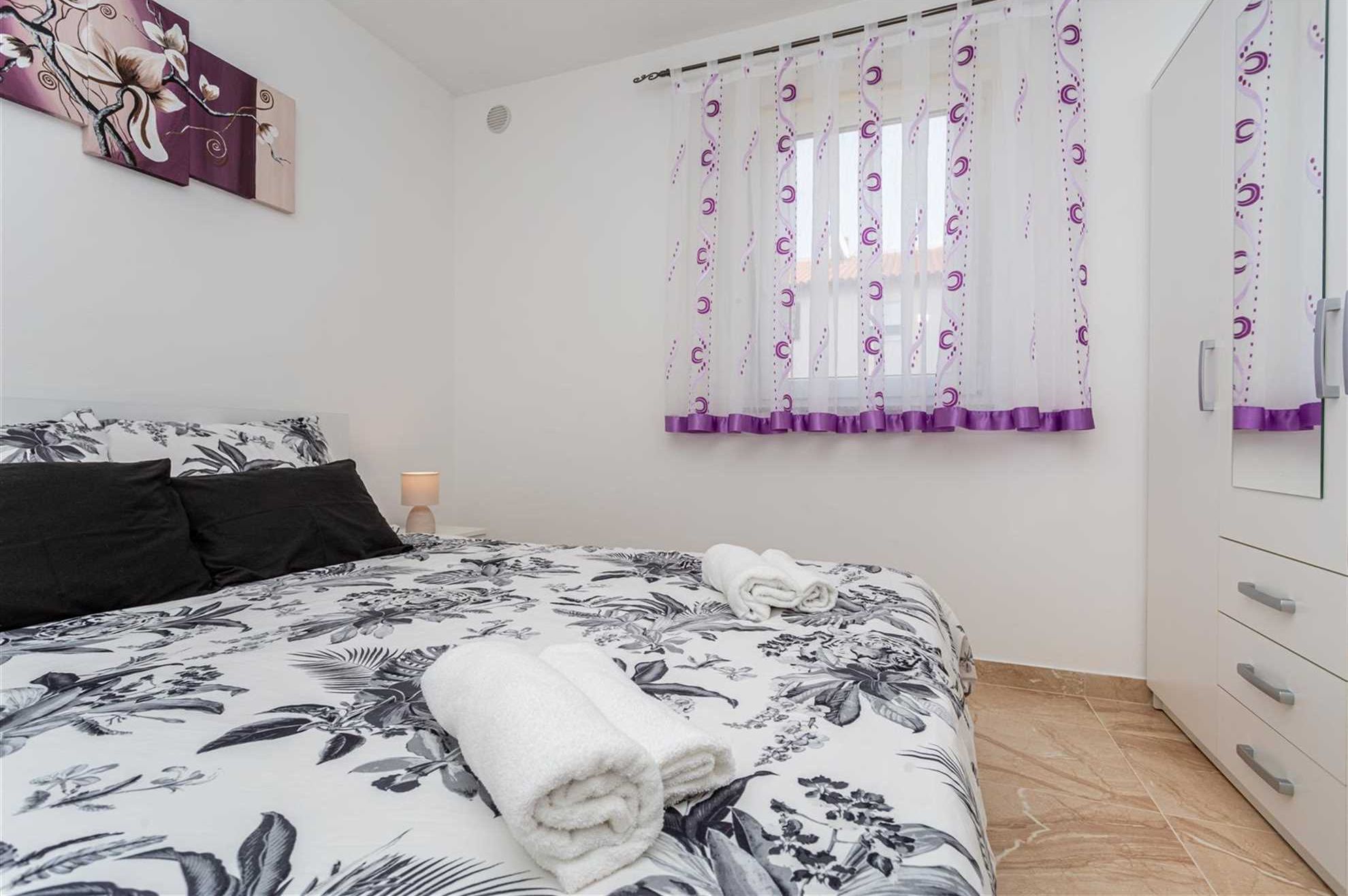 A bedroom with a bed, a nightstand, a lamp, a window with purple curtains, and a mirror.