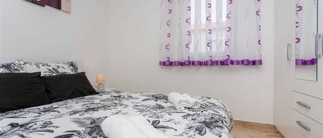 A bedroom with a bed, a nightstand, a lamp, a window with purple curtains, and a mirror.