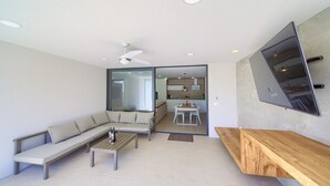 Una sala de estar moderna con un sofá, una mesita de centro y un banco de madera. Se observa un ventilador de techo y una cocina a través de una puerta abierta.