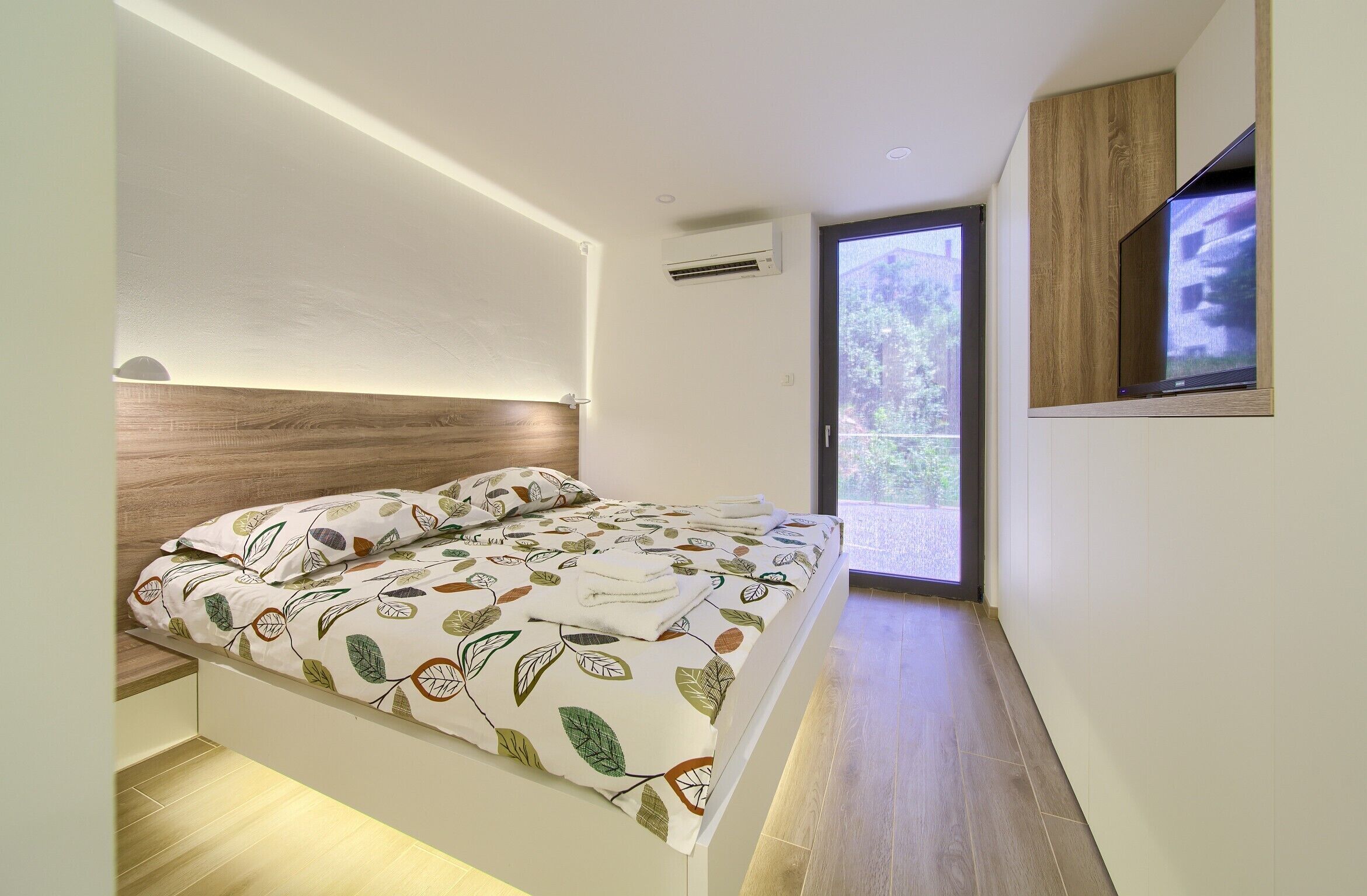 A modern bedroom with a bed, a TV mounted on the wall, and a large window.