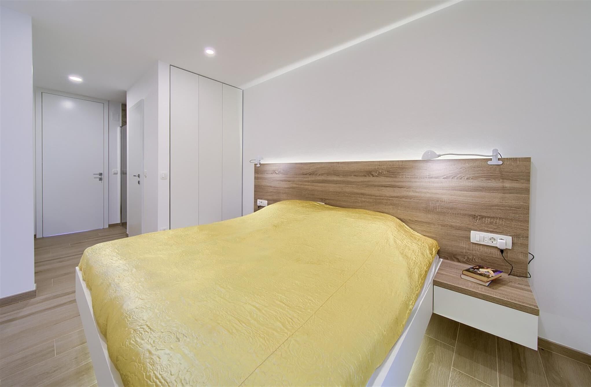 A modern bedroom with a bed covered in a yellow blanket, a wooden headboard, a bedside table with a book, and a white wardrobe.
