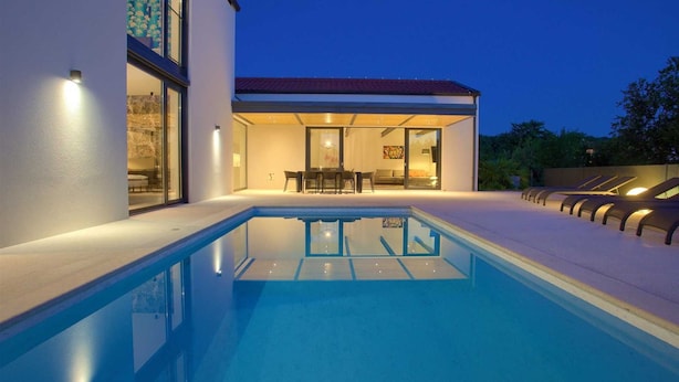 Una casa moderna con piscina, un patio con mobiliario exterior y un interior bien iluminado.