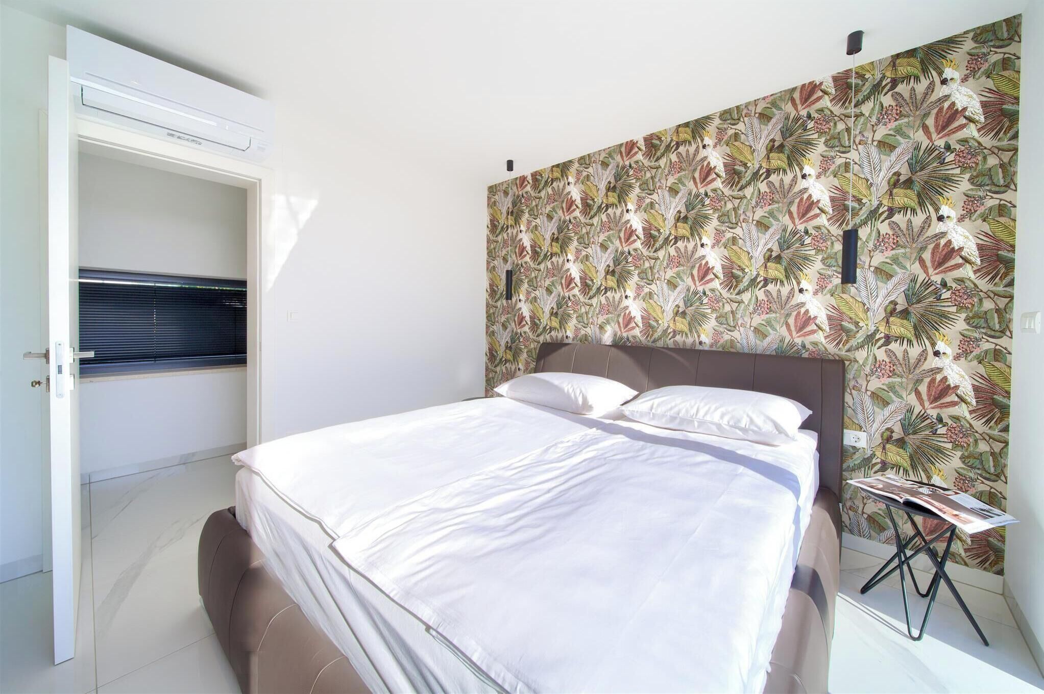 A bedroom with a bed, a window with blinds, a wall with a tropical leaf pattern, and a small table with a magazine.