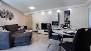 Un soggiorno moderno con un divano grigio, un'area pranzo con un tavolo rotondo apparecchiato per quattro persone e un grande specchio che riflette l'ambiente.