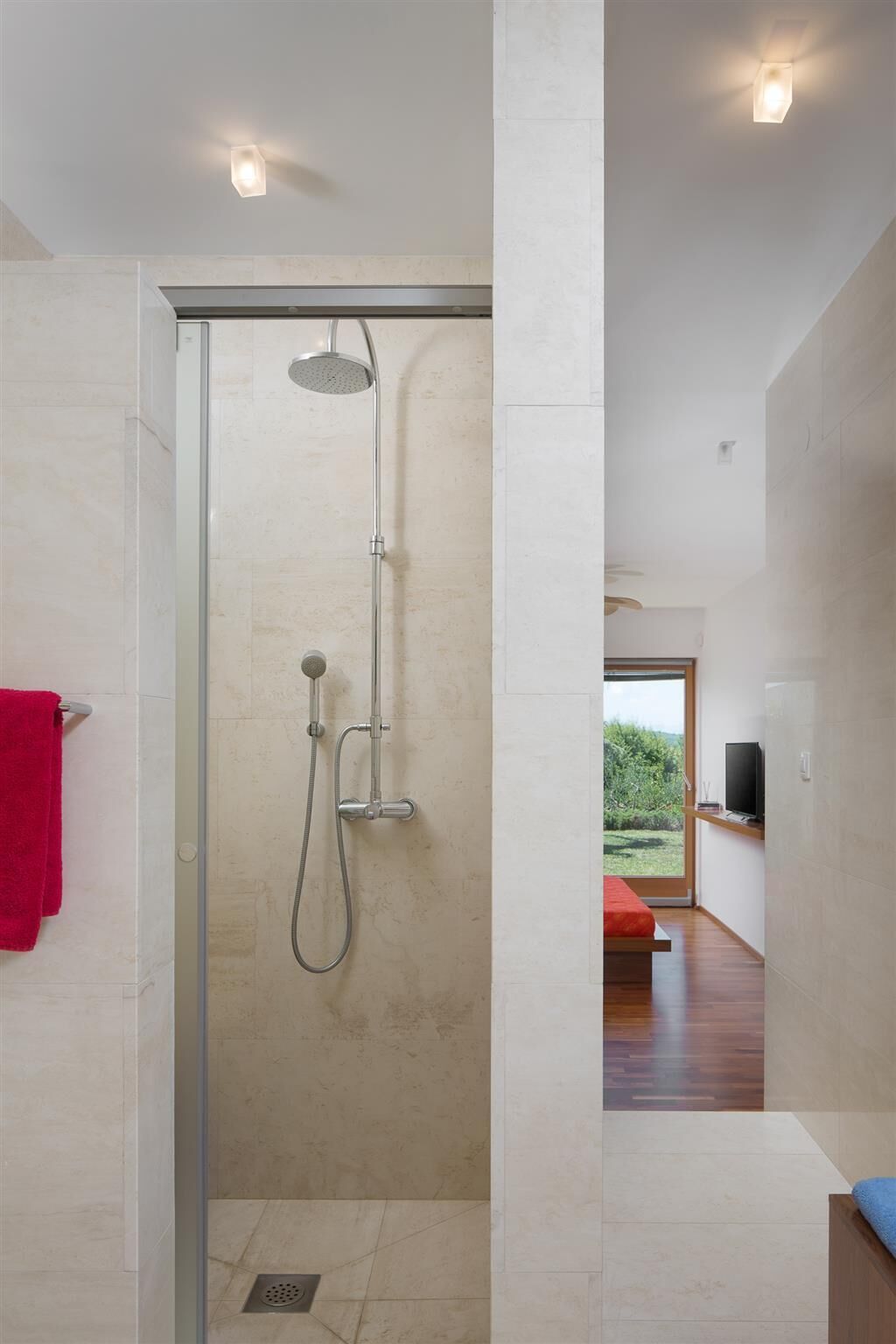 A modern bathroom with a glass-enclosed shower, a visible towel rack, and a view of a wooden floor and a red bench in the adjacent room.