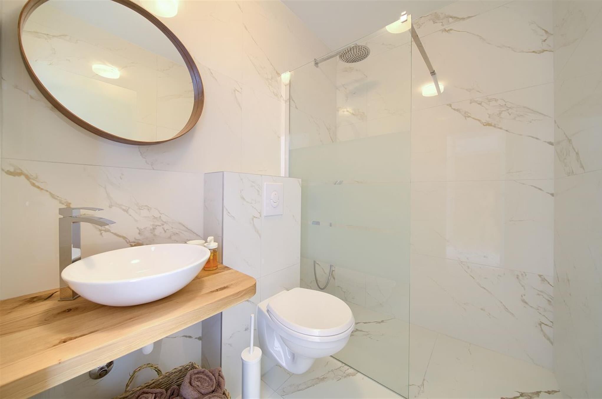 A modern bathroom with a marble-tiled wall, a wooden vanity with a white sink, a toilet, and a glass-enclosed shower.