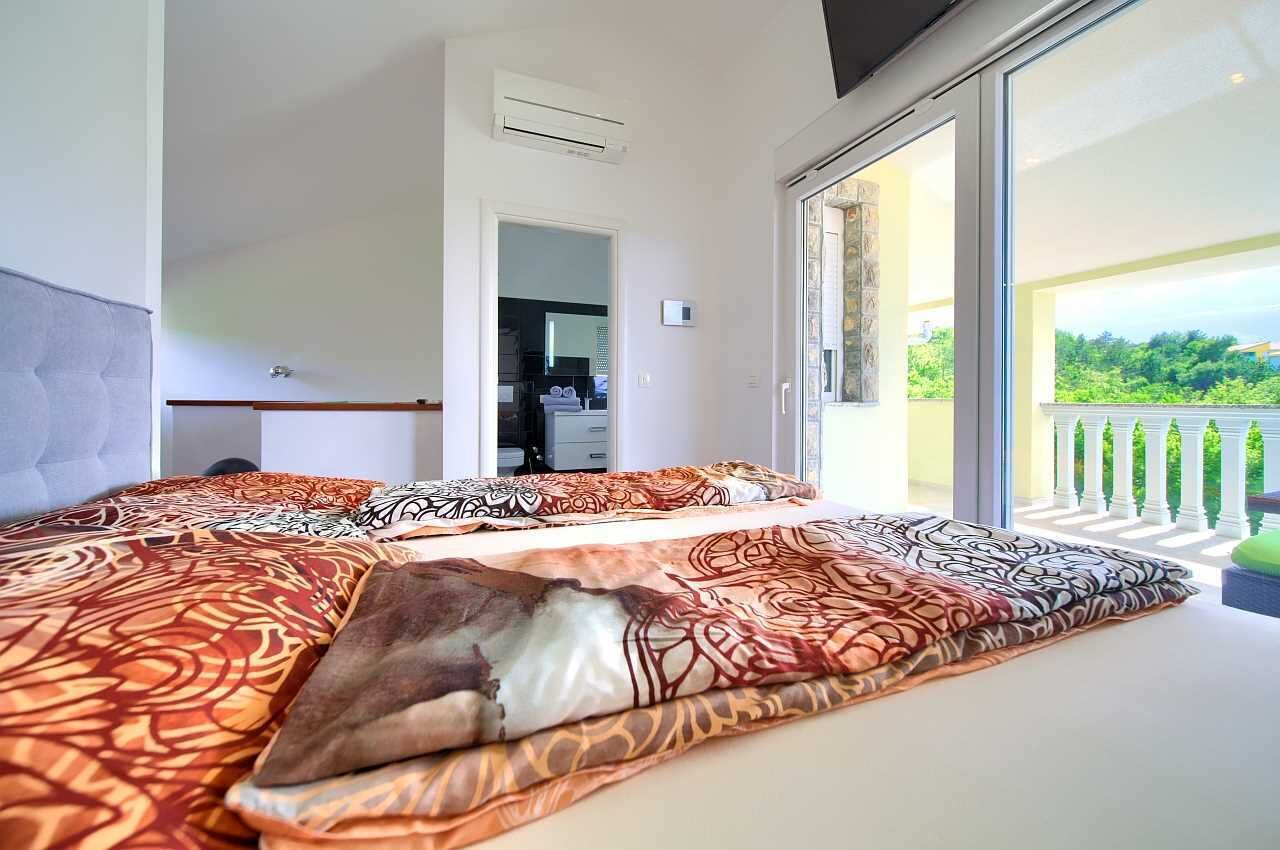 A bedroom with a large bed, a sliding glass door leading to a balcony with a view of greenery.