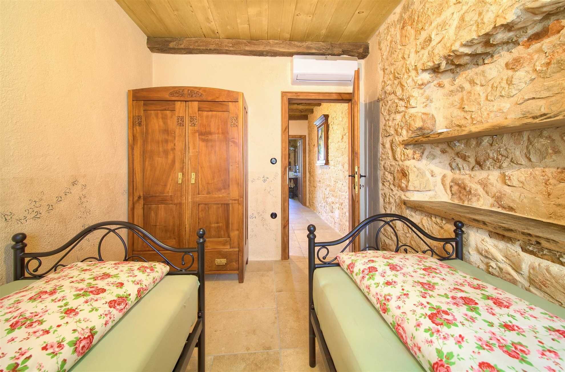 Un pasillo estrecho con dos camas de madera, cada una con cubrecama floral, que conduce a una pared de piedra y una puerta de madera.