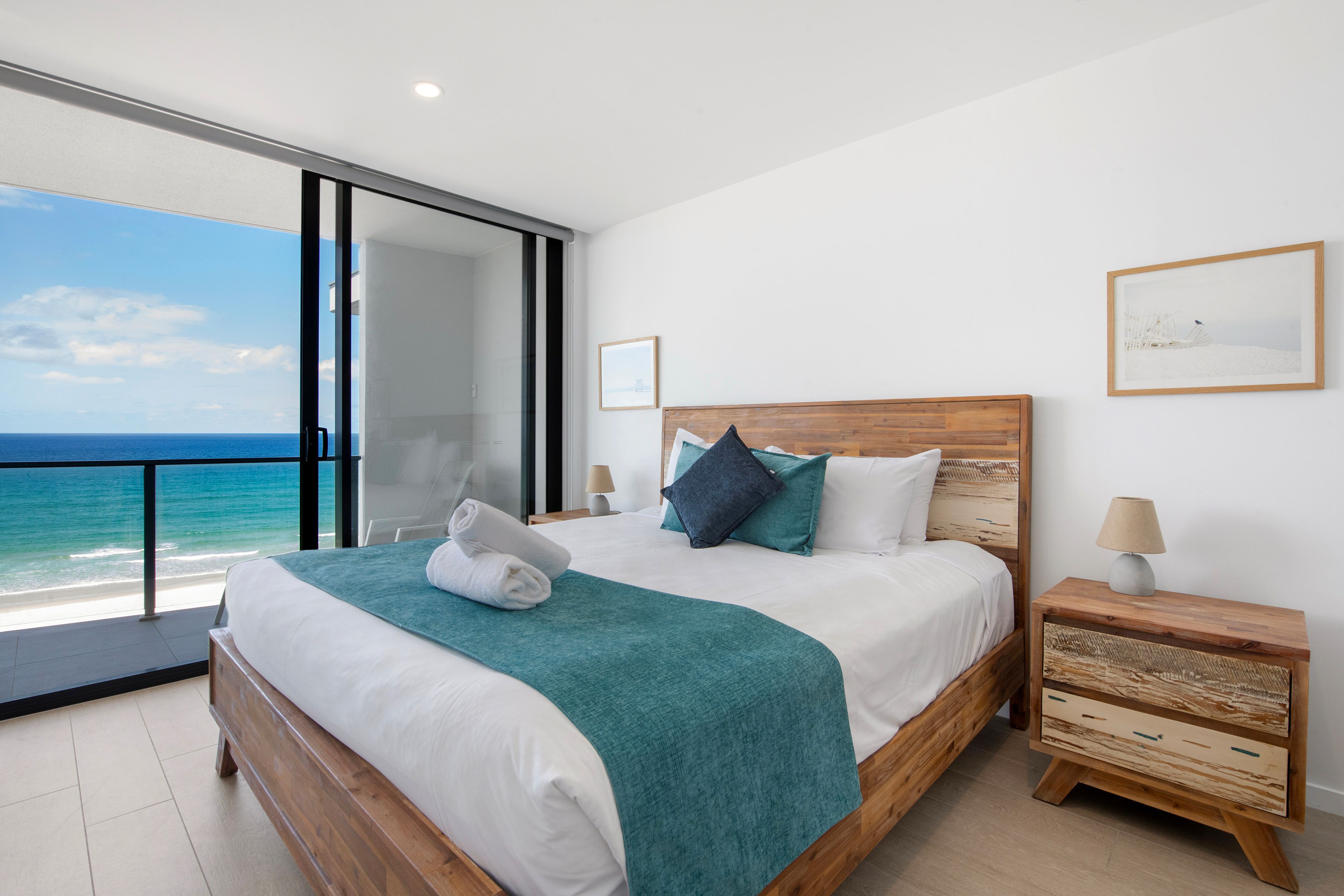A bedroom with a large bed, a nightstand, a wooden headboard, a view of the ocean, and a glass door leading to a balcony.