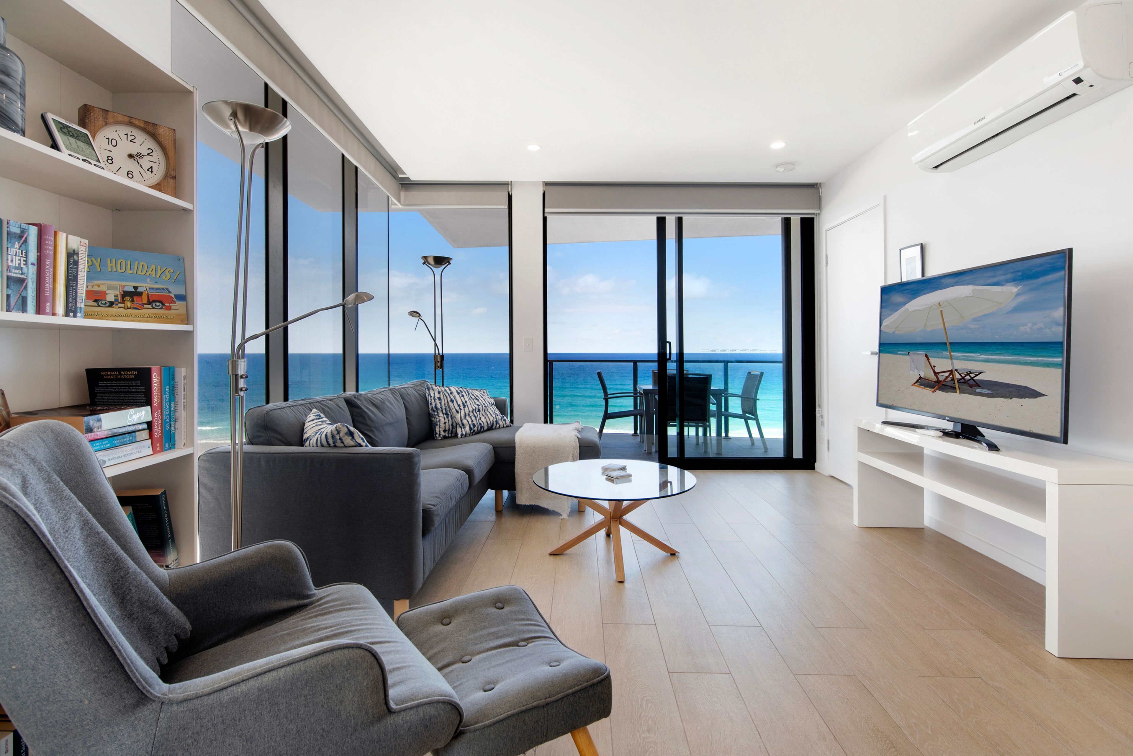 A modern living room with a sofa, a coffee table, and a television. There is a bookshelf with books and a clock. The room has large windows with a view of the ocean.