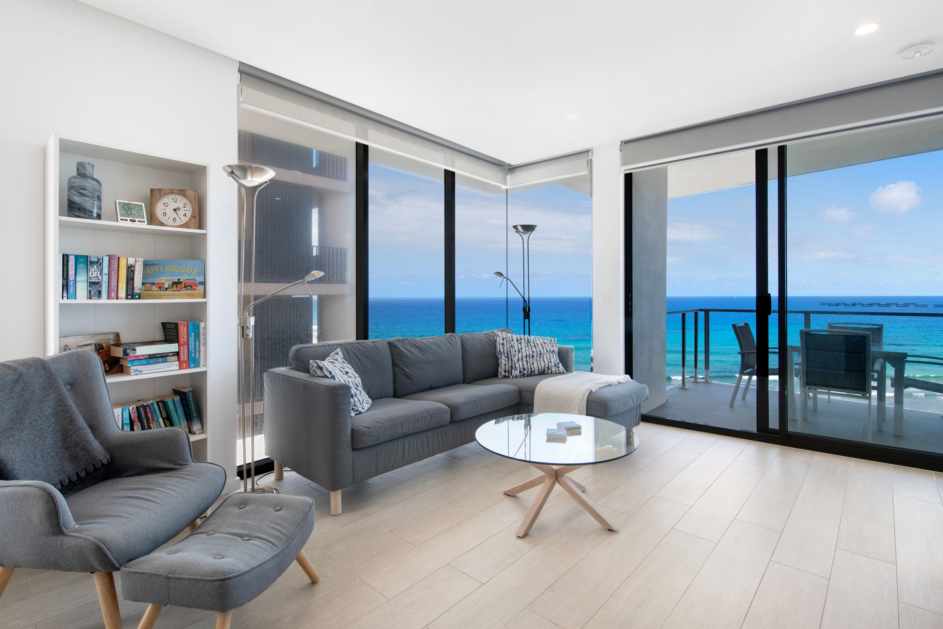 A modern living room with a large window overlooking the sea, a sofa, a coffee table, and a bookshelf.