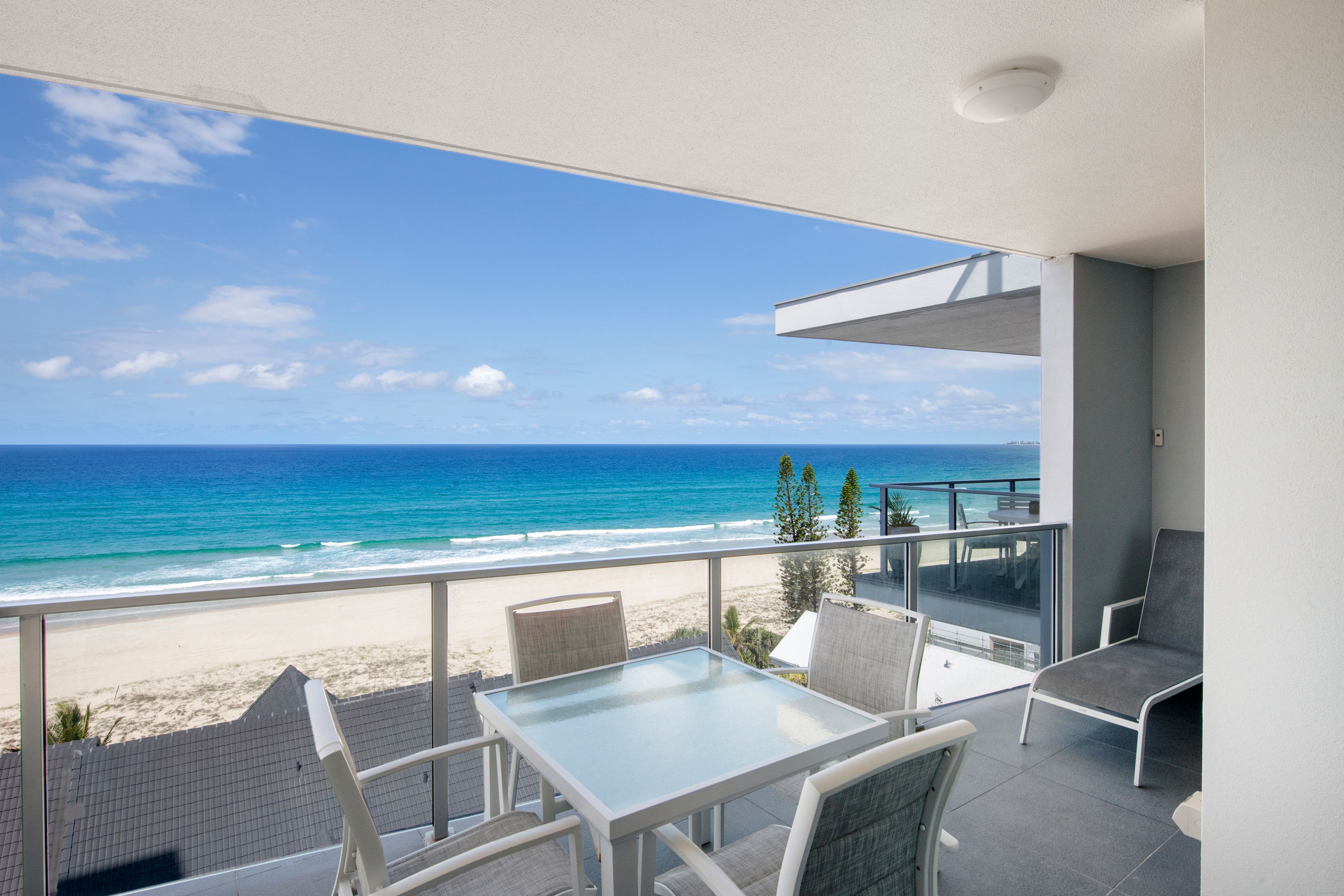 A balcony with a glass table and chairs, overlooking a beach and ocean.