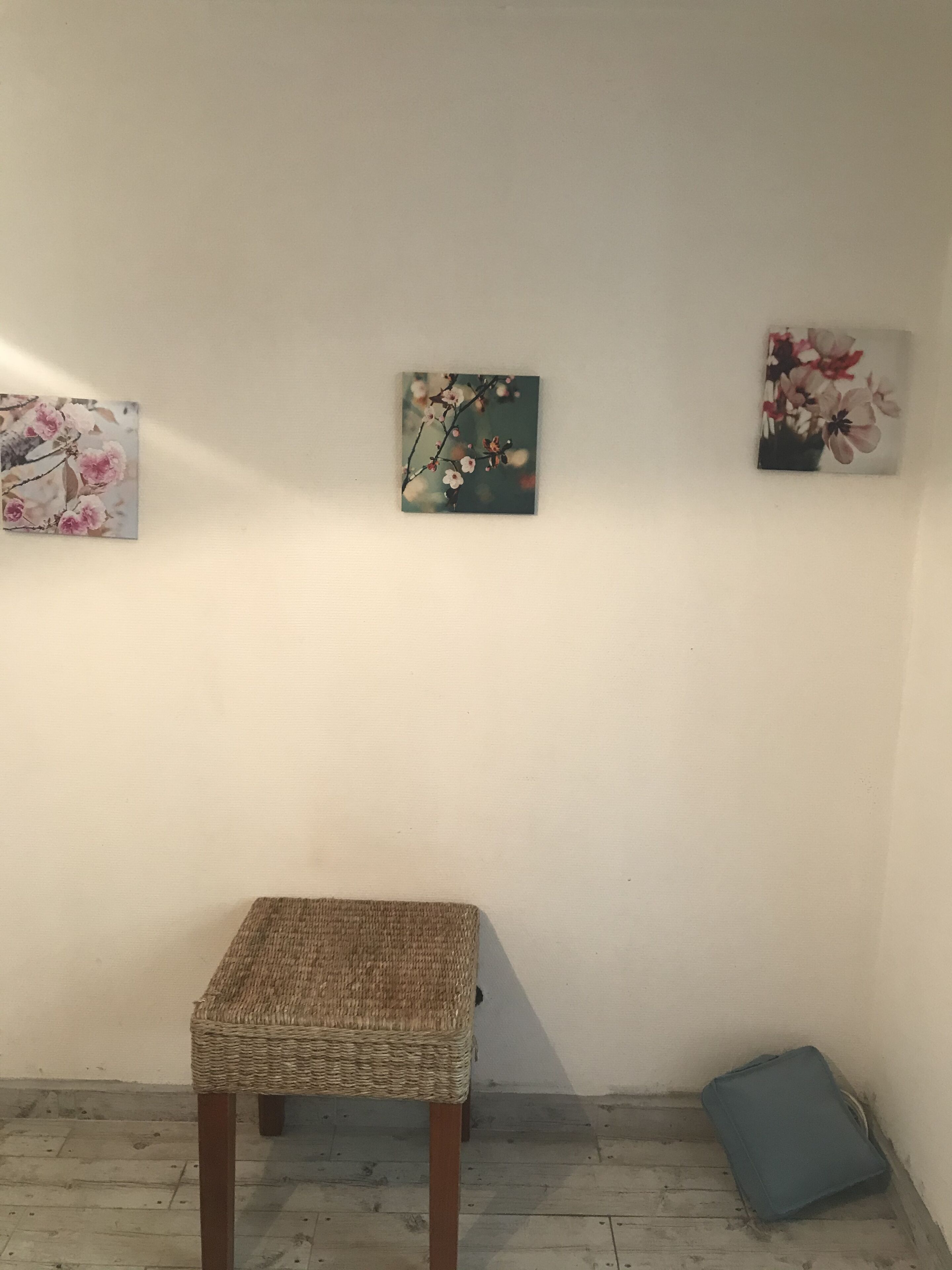 A room with a wicker chair, a small table, and three wall-mounted paintings.