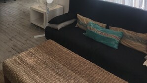 Ein modernes Wohnzimmer mit einer schwarzen Couch, einem gewebten Teppich und einem Standventilator. An der Wand hängen gerahmte Bilder und ein Fenster mit Vorhängen.