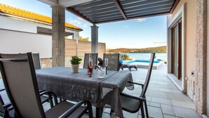 An outdoor patio with a table and chairs, a view of a swimming pool, and a stone pillar. - Villa Ema with Seaview, swimming pool, garage, BBQ & playroom (Čižići (Krk))