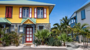 Une maison colorée à deux étages, avec une porte rouge et de nombreuses fenêtres, entourée de plantes tropicales et surplombant un ciel d’un bleu limpide.