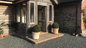 A wooden house with a gabled roof, a porch with two potted plants, and a gravel driveway.