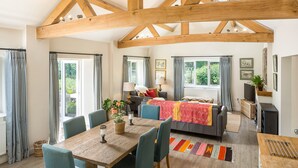 A spacious living area with a wooden ceiling, a dining table with chairs, a sofa, and a window with curtains.