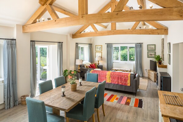 A spacious living area with a wooden ceiling, a dining table with chairs, a sofa, and a window with curtains.