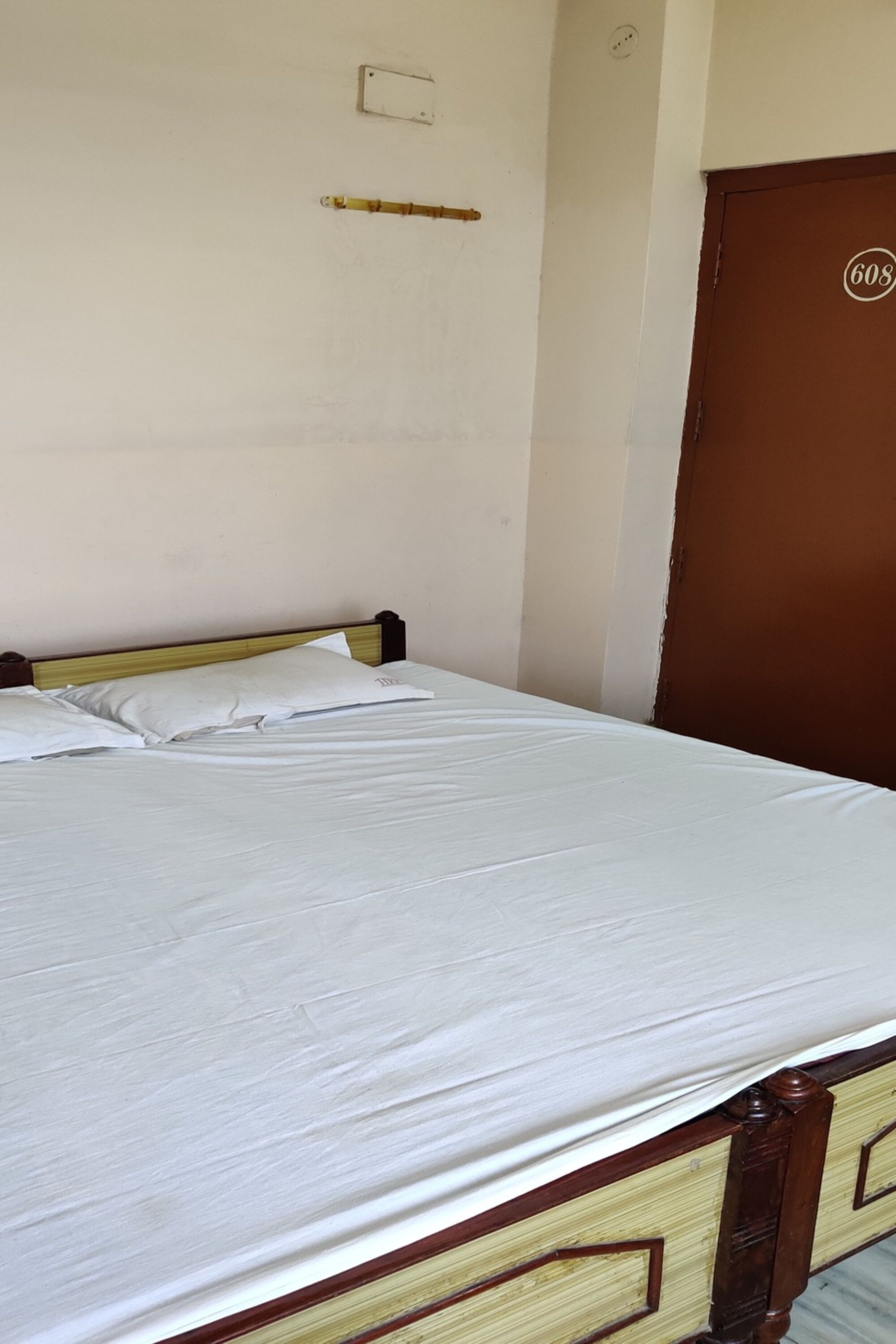 A single bed with a wooden frame and a white mattress in a room with a plain wall and a brown door.
