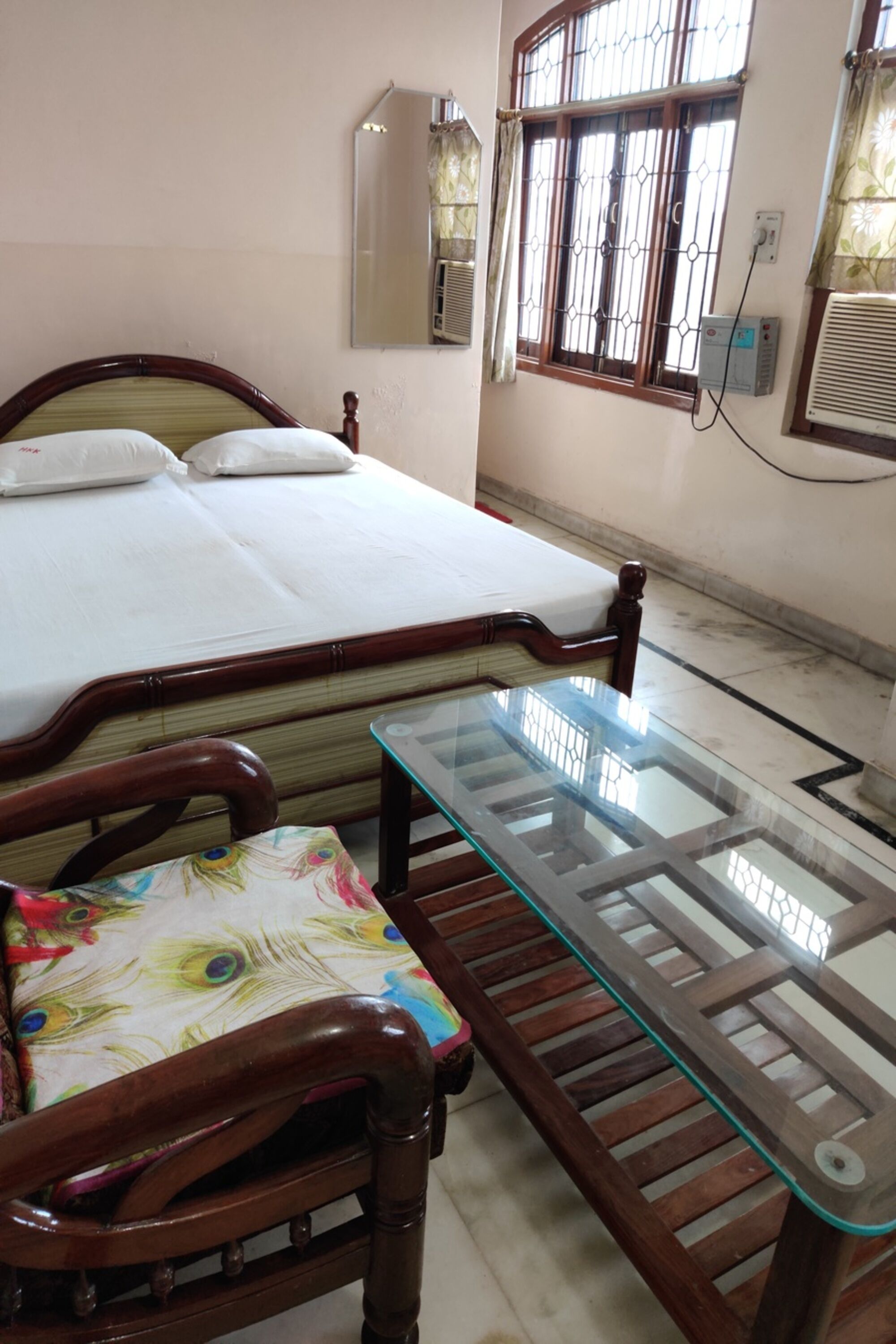 A bedroom with a bed, a wooden chair, a glass-top table, a mirror, and a window with curtains.