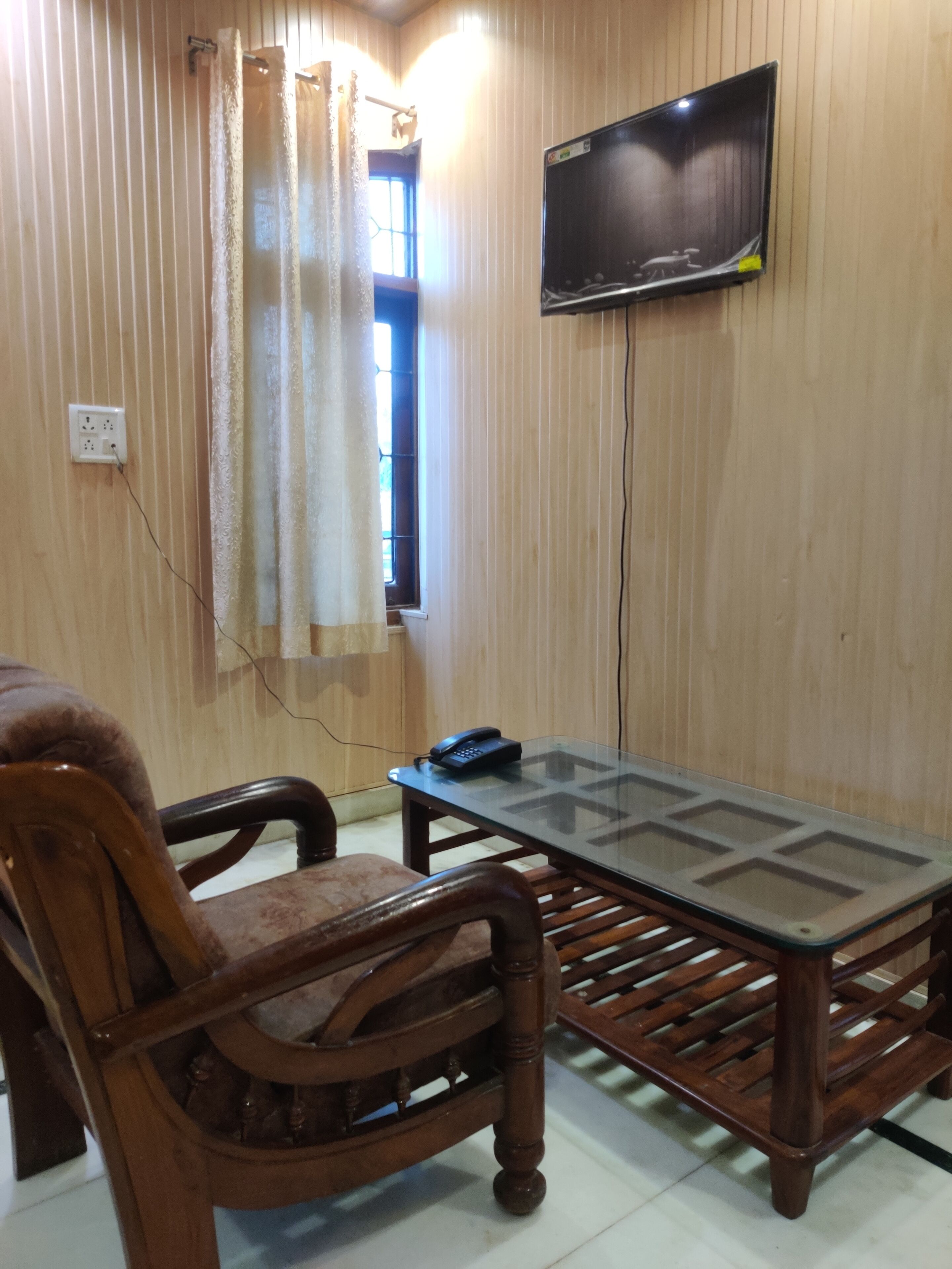 Une pièce avec un mur lambrissé en bois, un téléviseur à écran plat fixé au-dessus d’une table en verre, un téléphone sur la table et une fenêtre avec des rideaux.