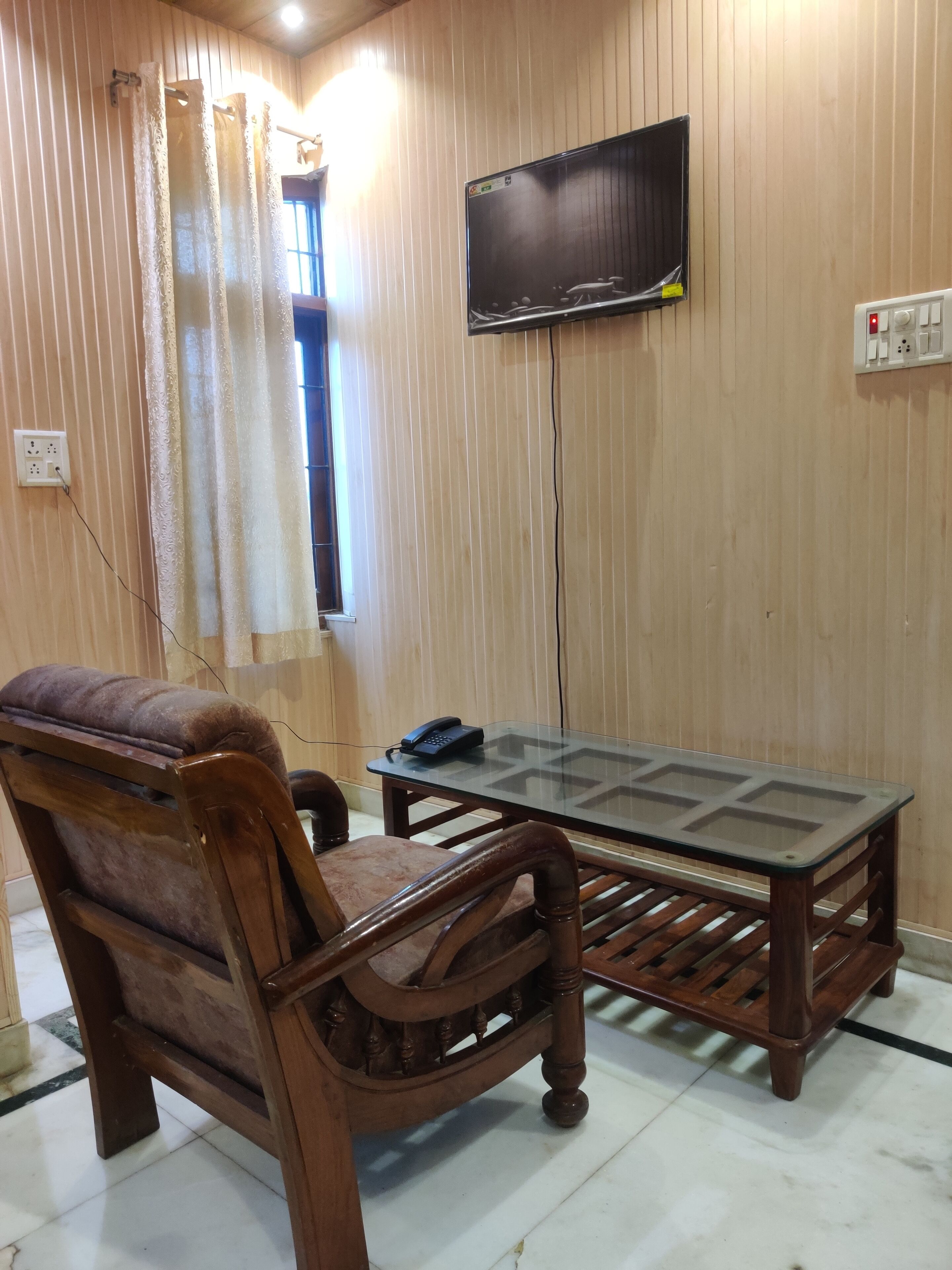Um quarto com parede revestida de madeira, televisão montada acima de uma mesa de vidro, uma poltrona acolchoada e uma janela com cortinas.