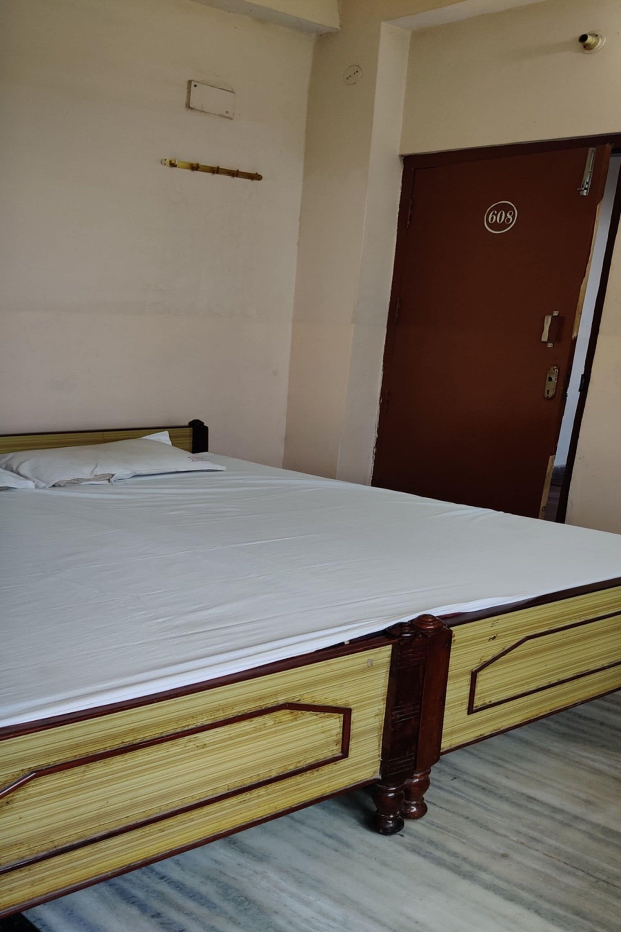 Una cama individual con estructura y cabecera de madera, una puerta marrón con placa de número y una pequeña estantería sobre la cama.
