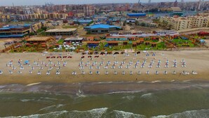A beach with rows of white sun loungers and umbrellas, a playground, and a view of a cityscape in the background. - Jewel Port Said Hotel (Port Said)