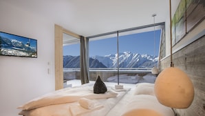 Un dormitorio con un ventanal que ofrece vistas a la montaña, una cama con ropa de cama blanca, una almohada y una lámpara colgante.
