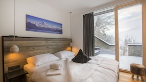 Un dormitorio moderno con cabecera de madera, una cama grande con ropa blanca, un cuadro de un paisaje montañoso, vista a un área nevada a través de un ventanal y un taburete de madera.
