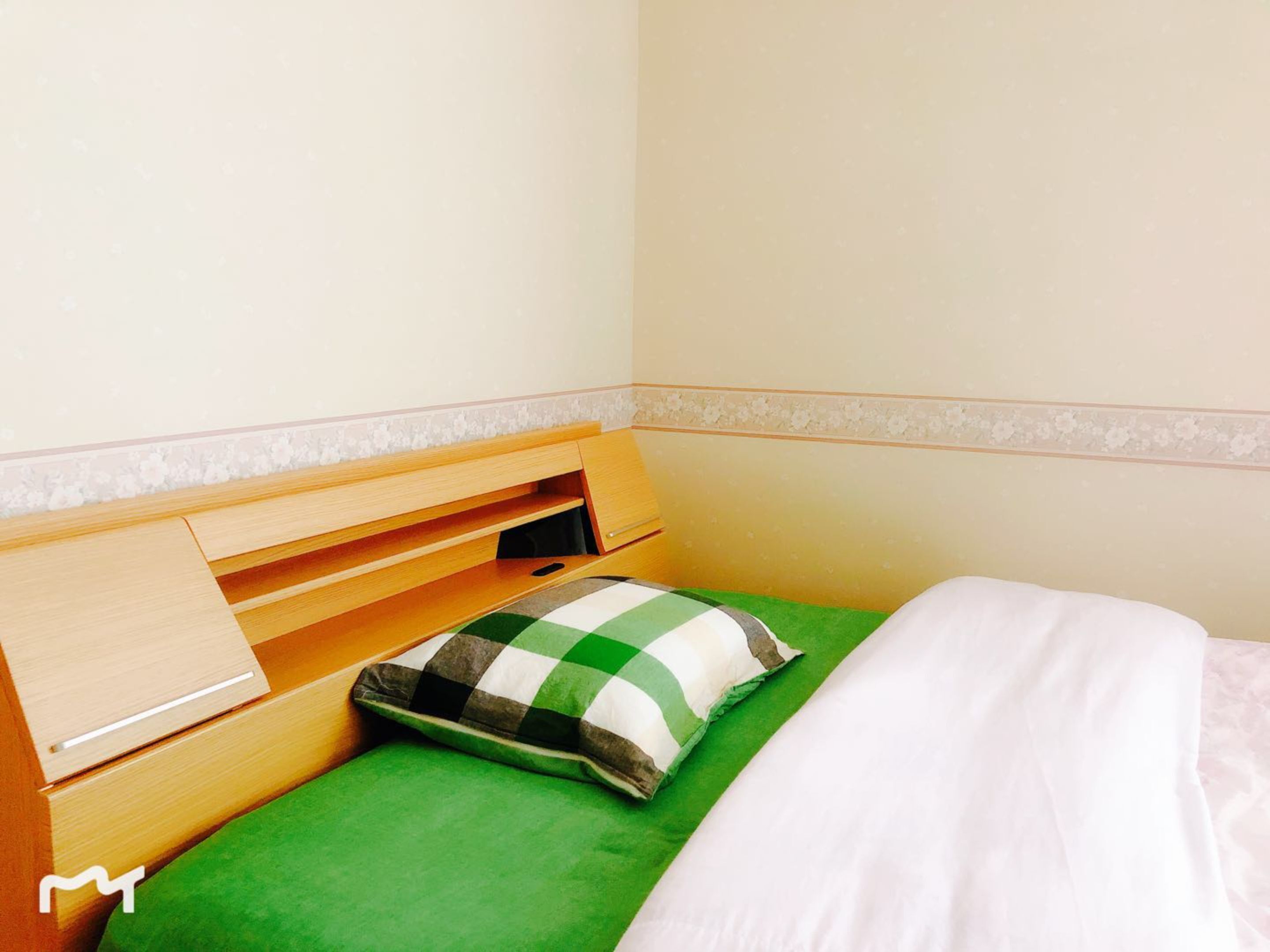 A small, neatly made bed with a green bedspread and a checkered pillow, a wooden headboard with a drawer, and a plain wall with a floral border.