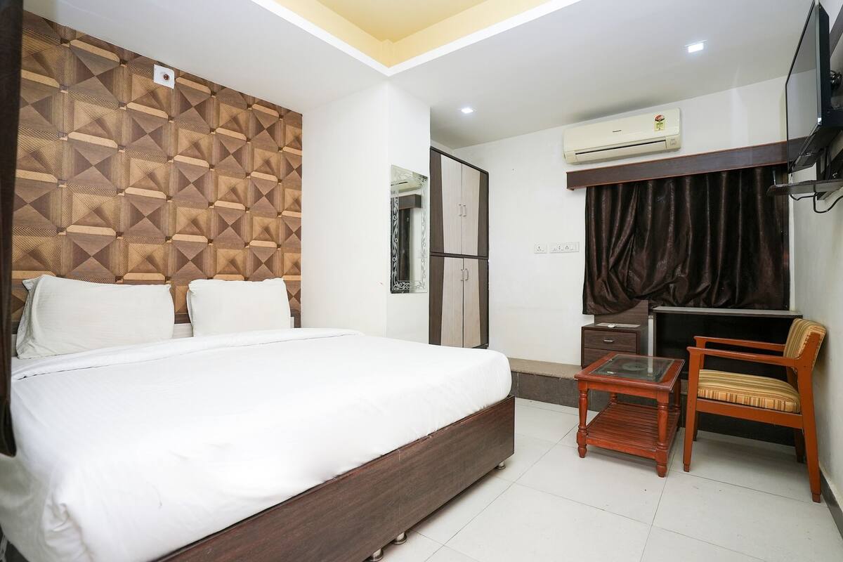 Quarto com cama grande, cabeceira de madeira, mesinha, cadeira, aparelho de ar condicionado e televisão.