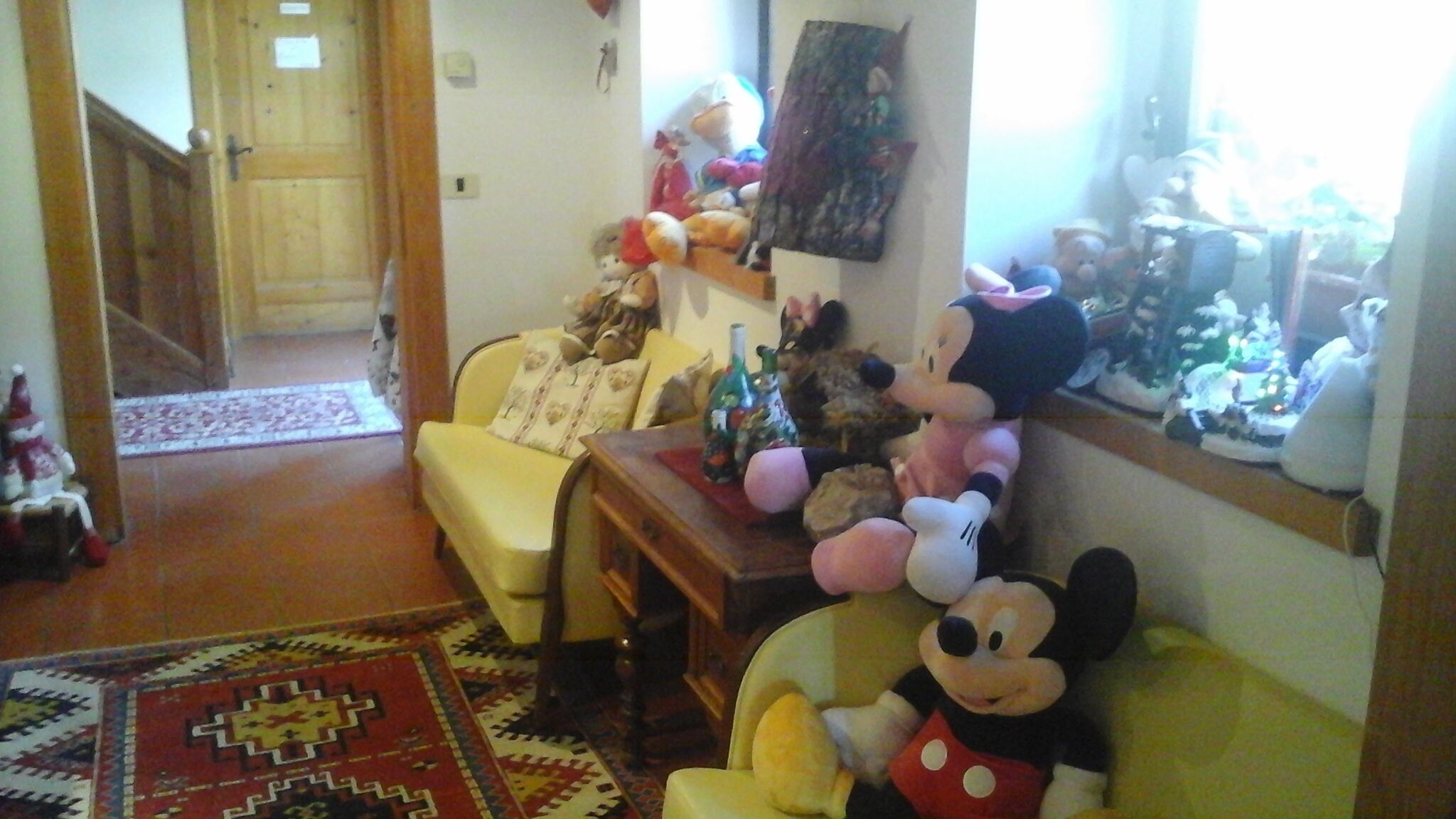 A living room with a yellow armchair, a table with a Mickey Mouse plush toy, and a shelf with various stuffed animals and figurines.