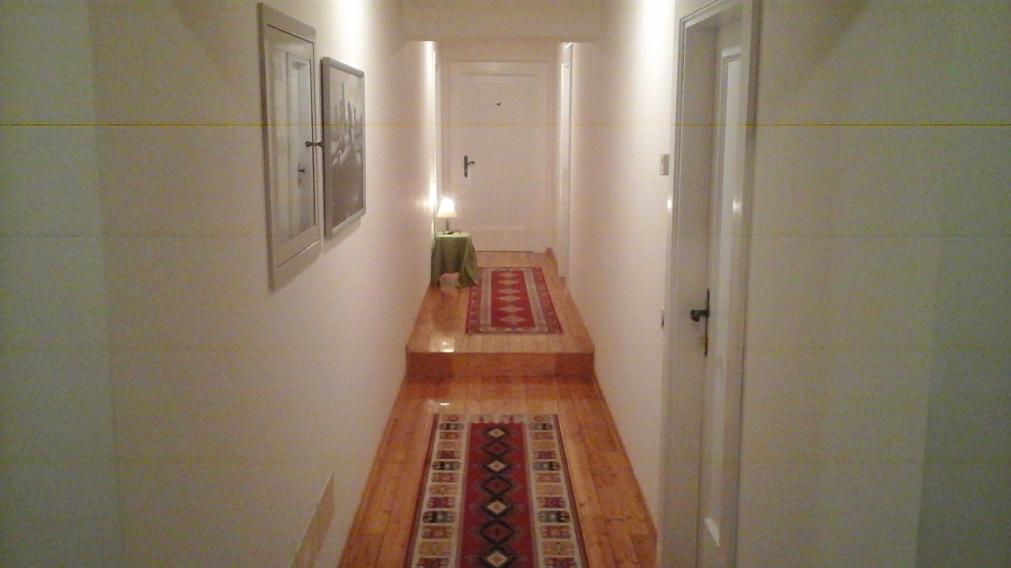 A narrow hallway with a wooden floor, a red patterned rug, and a small table with a lamp.